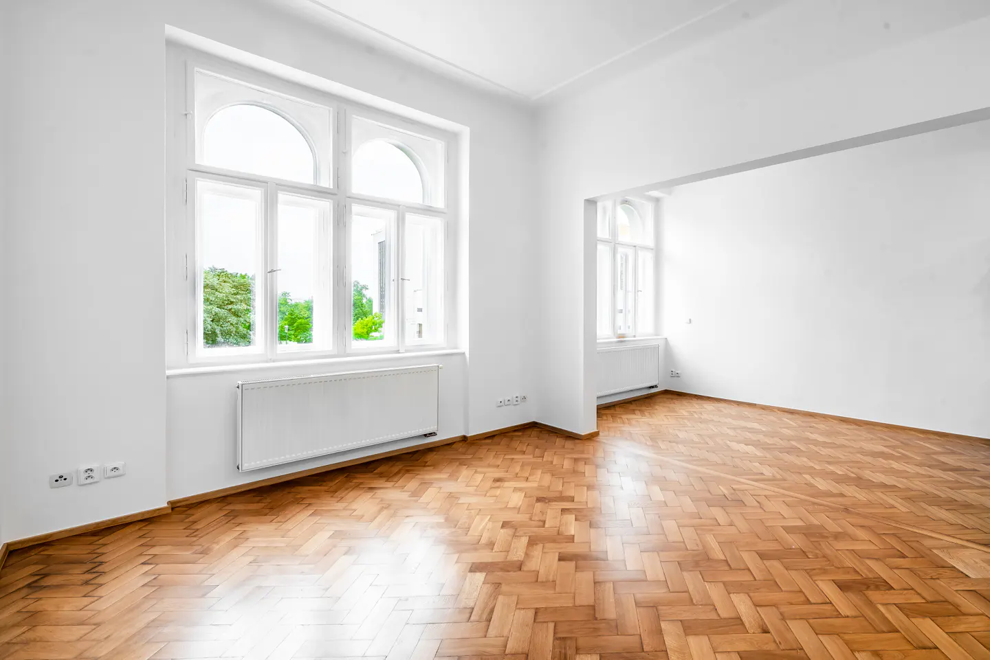 Bright, empty room with herringbone wood floors and white walls. Large arched windows let in natural light. A radiator sits beneath the window.