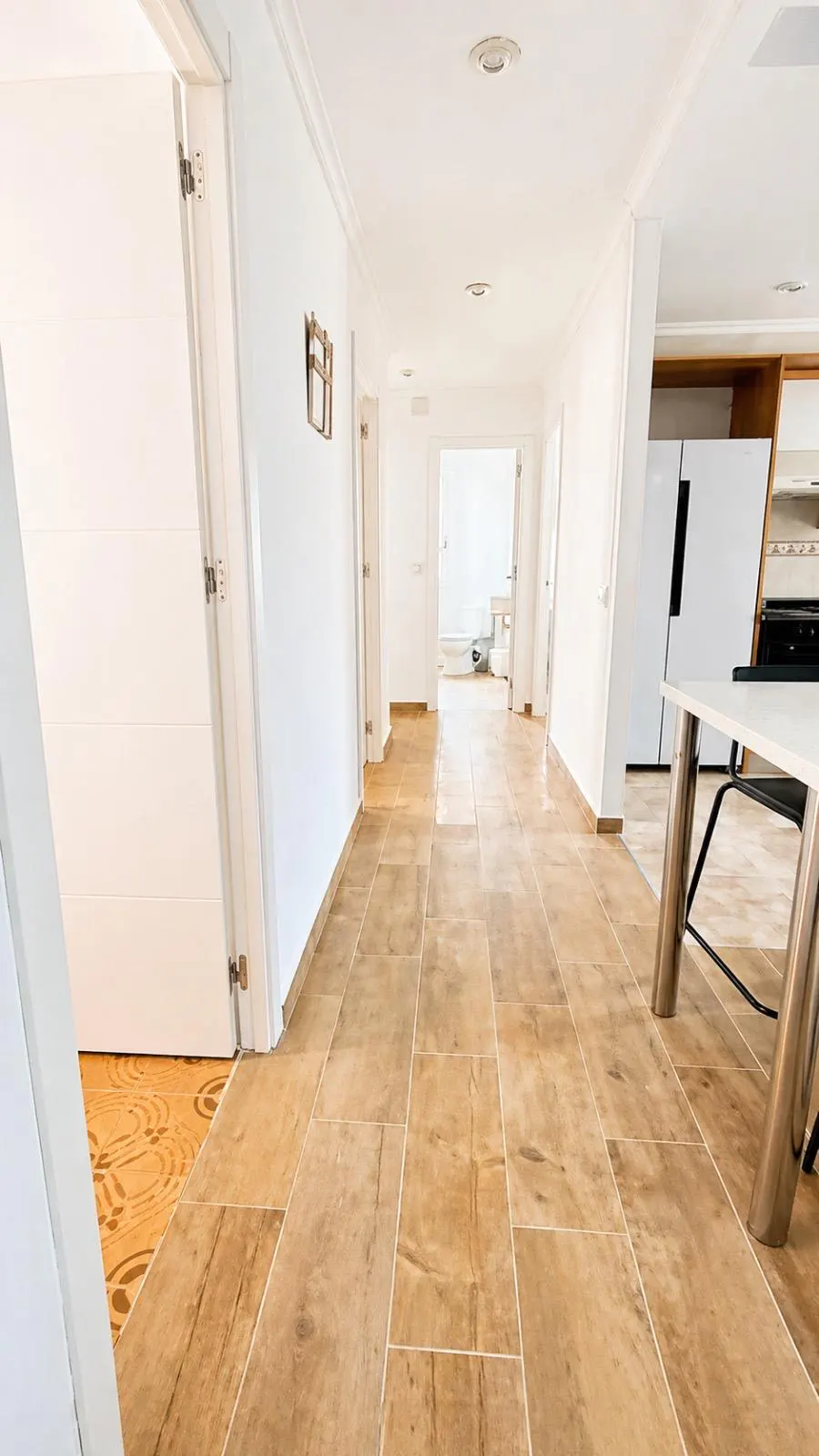Bright hallway with wood-look tile floors, white walls and doors. A kitchen is visible at the end of the hall.