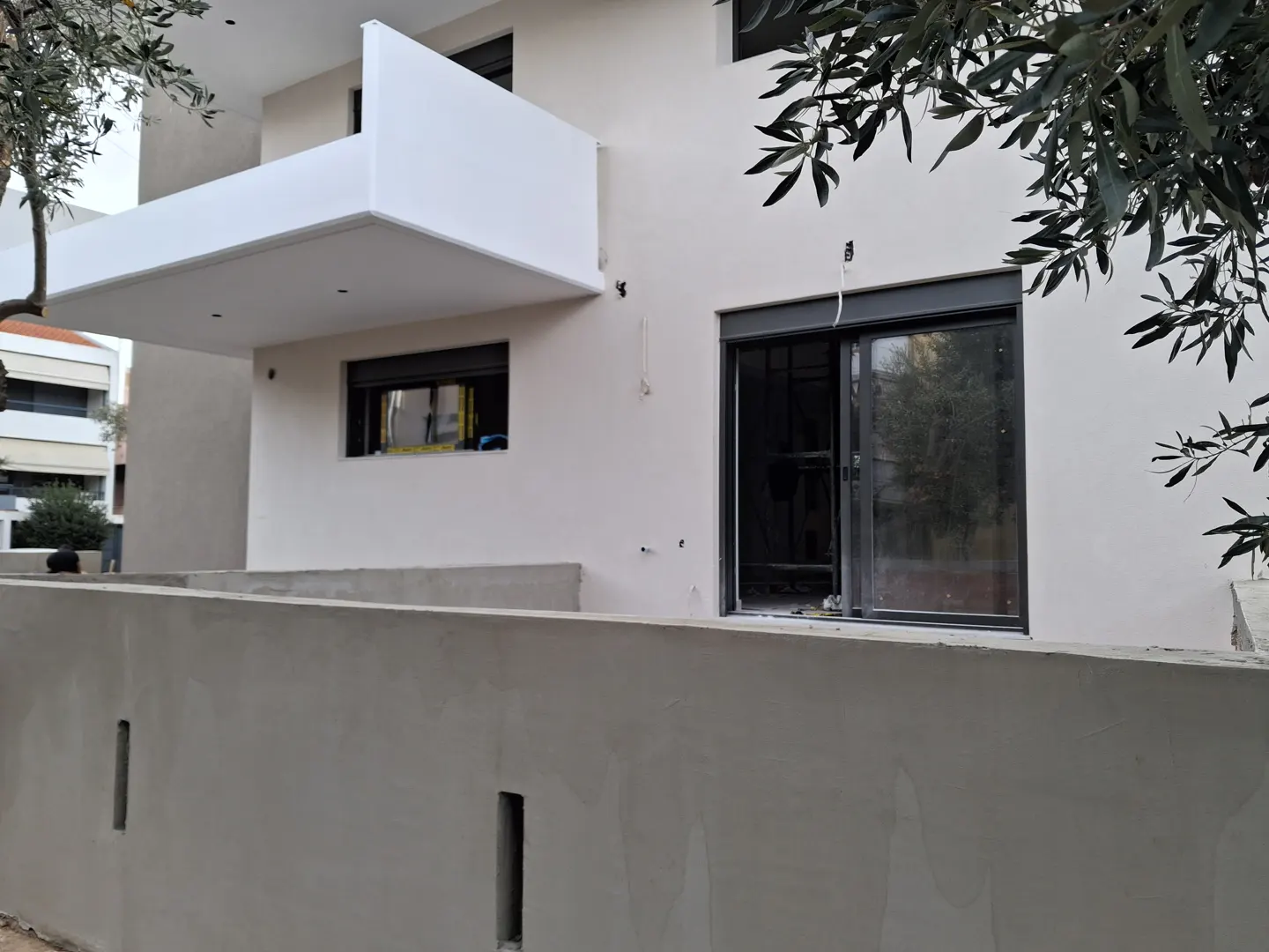 Modern beige house exterior under construction with a white balcony, dark window frames, and a concrete perimeter wall.