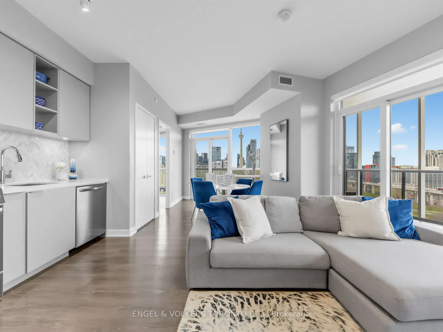 Modern open-concept condo with a grey sectional, kitchen, and floor-to-ceiling windows showing the Toronto skyline.