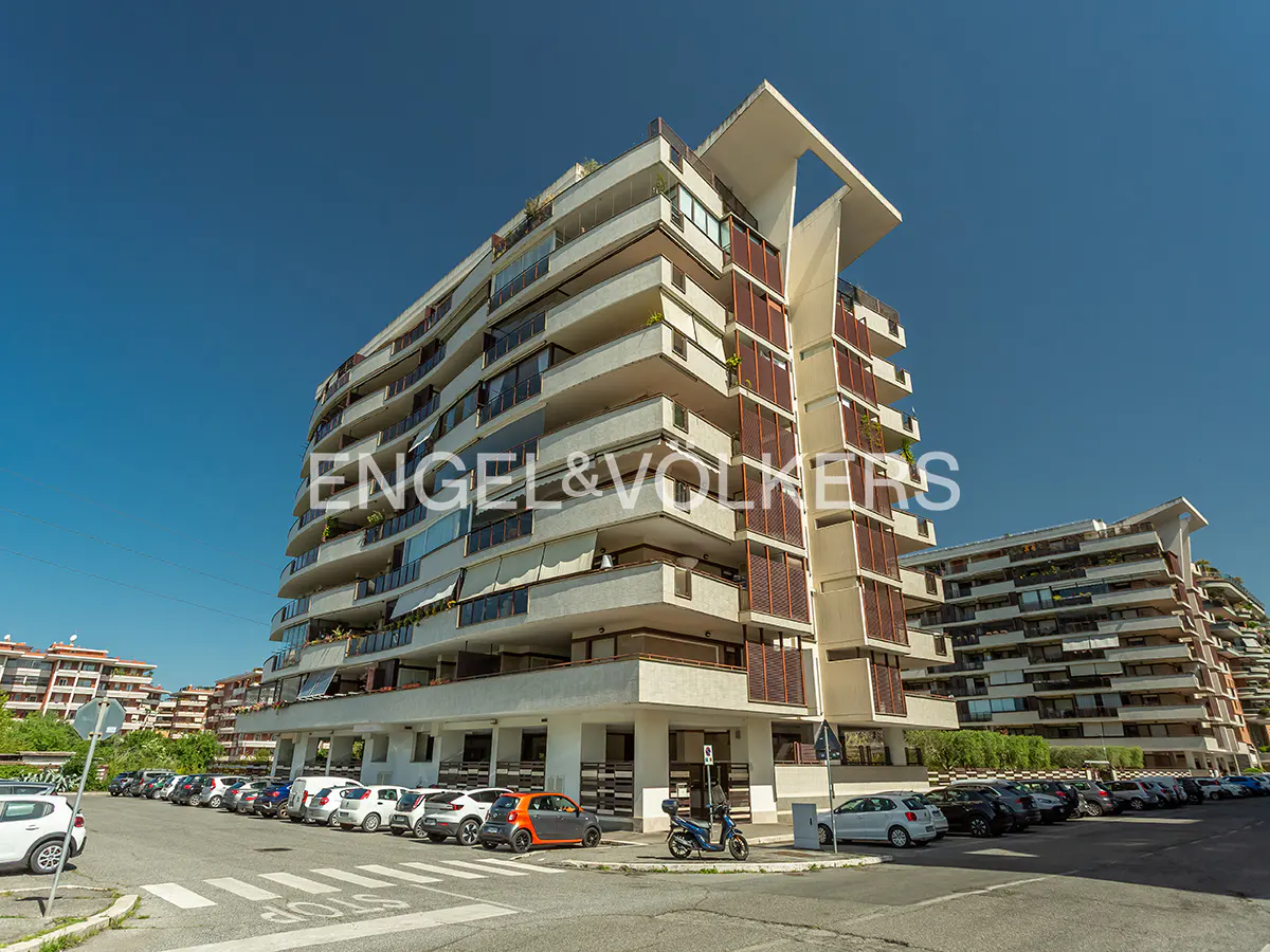 Exterior view of a modern, multi-story apartment building with white balconies and brown shutters under a clear blue sky. Cars parked in front.
