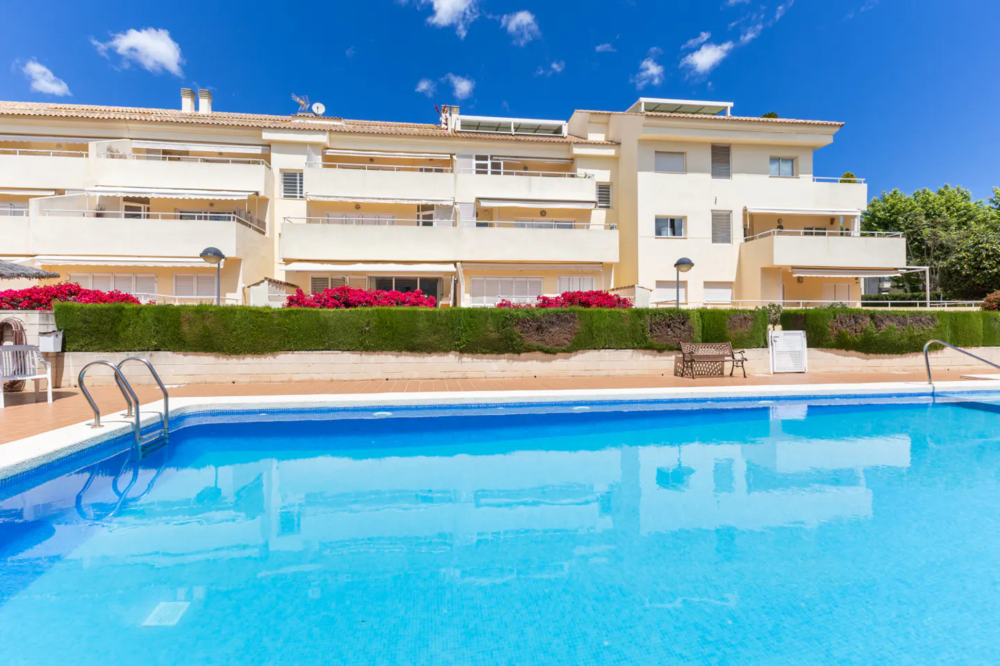 Exterior view of a multi-story, light yellow apartment building with a blue swimming pool in the foreground.