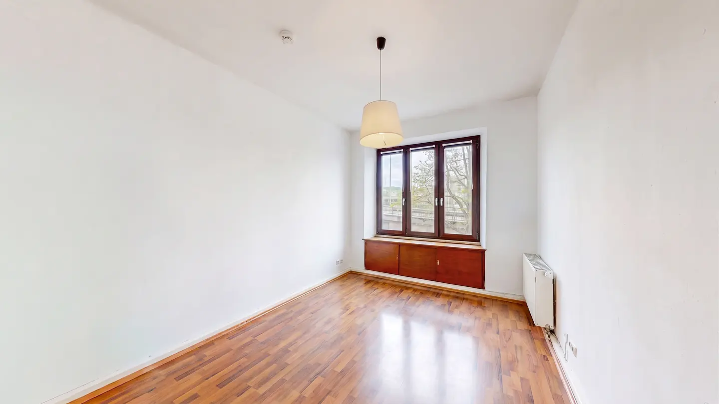 Bright, empty room with hardwood floors, white walls, and a window with a brown frame. A light fixture hangs from the ceiling.