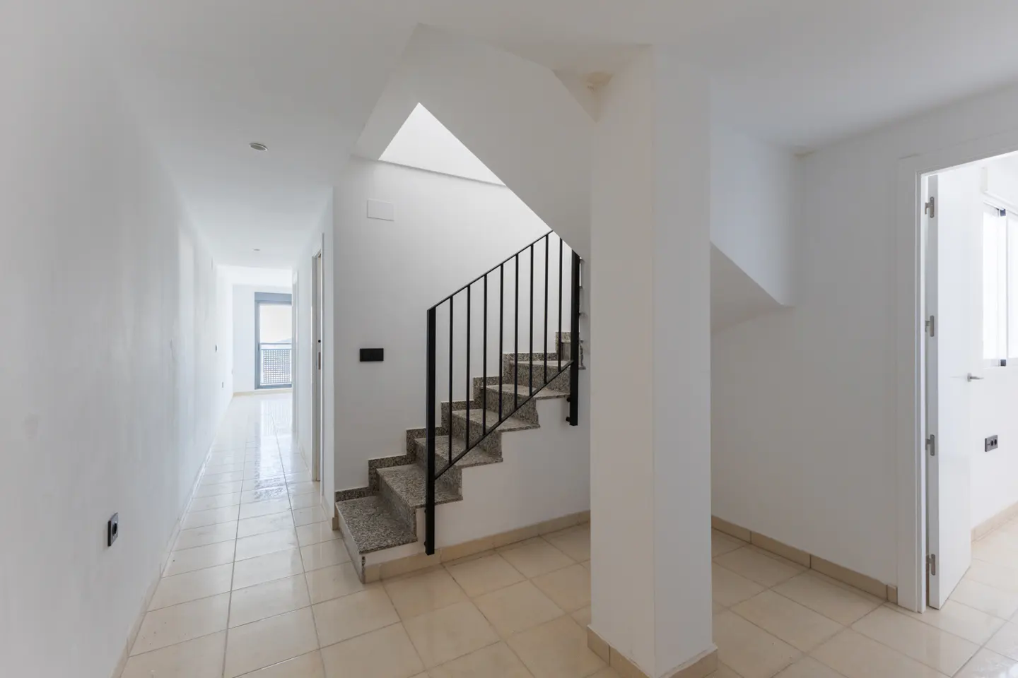 Interior view of a bright, white hallway with beige tile floors, a staircase with black railings, and an open doorway to another room.