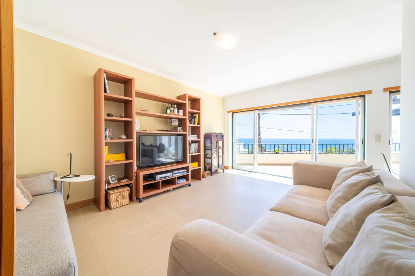 Bright living room with beige sofa, wooden shelves, and a TV. Sliding glass doors open to a balcony with ocean views.