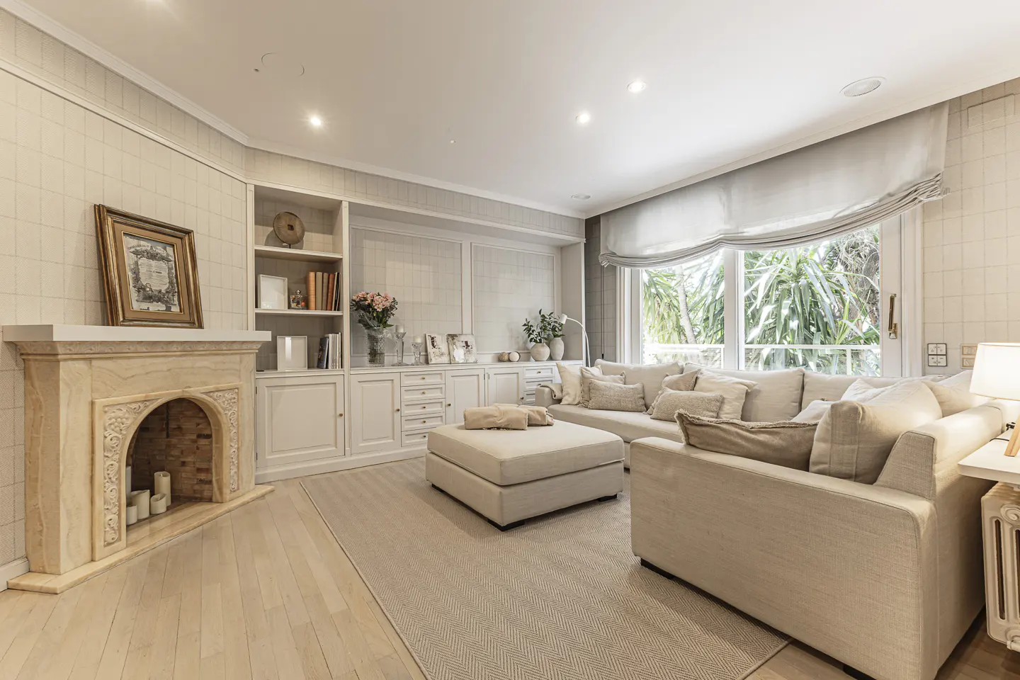 Living room with a beige sectional sofa, ottoman, fireplace, and built-in shelving. Large window with natural light.