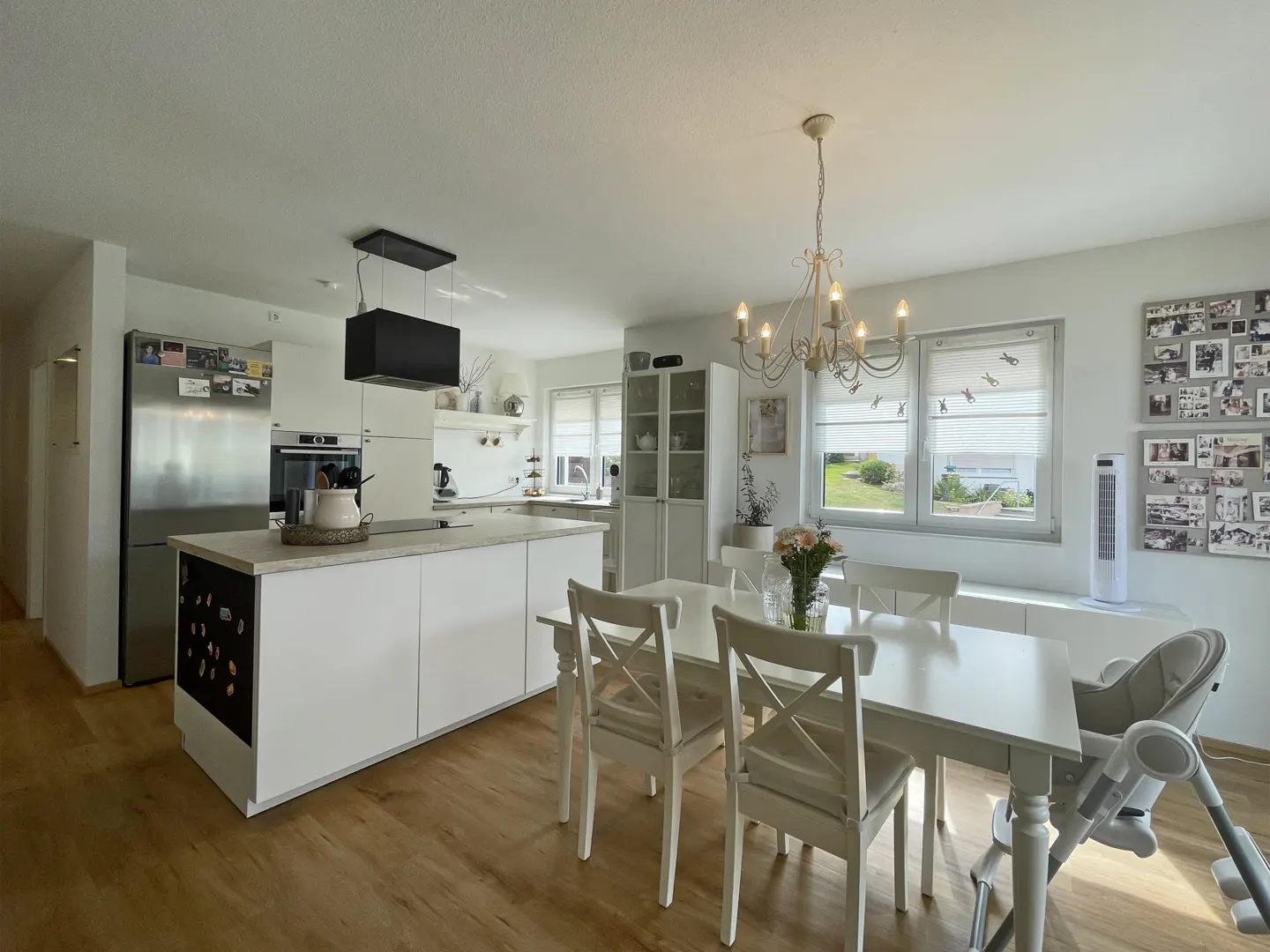 Bright, open-plan kitchen and dining area with white cabinets, island, and table. A chandelier hangs above the table, and windows offer a view of the outdoors.