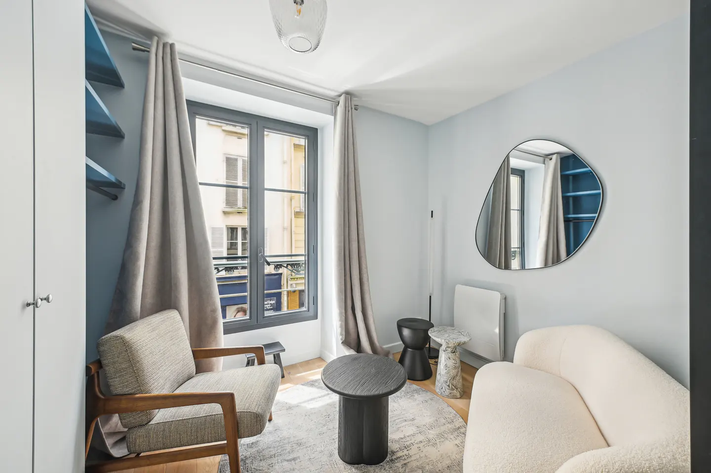 A bright living room with a gray chair, white sofa, and a round black table on a gray rug. A window with gray trim is draped with gray curtains.