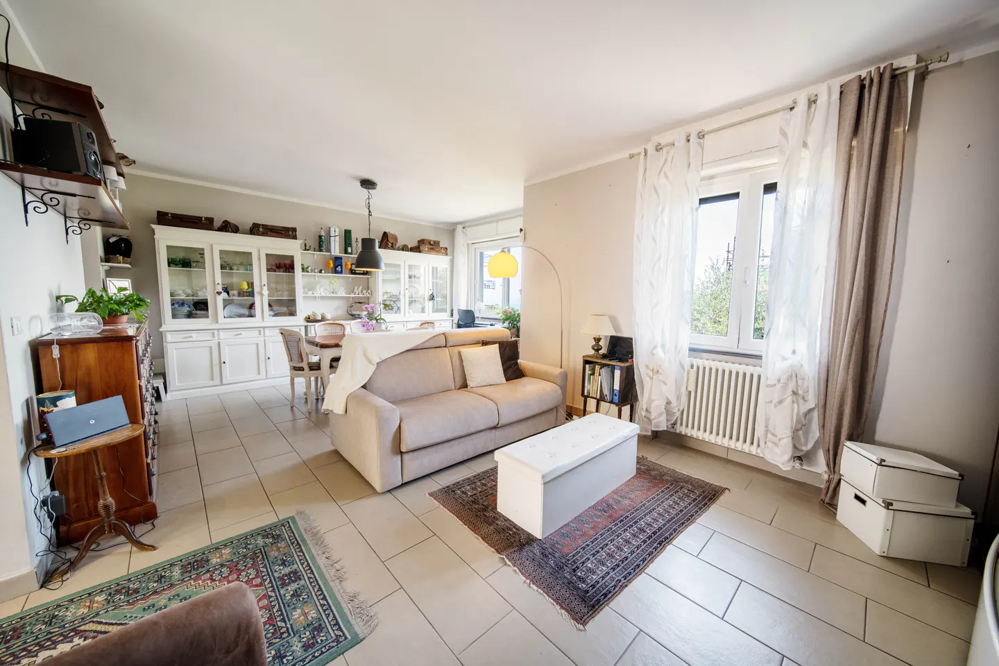 A bright living room with a beige sofa, white storage bench on a patterned rug, and white cabinets in the background.