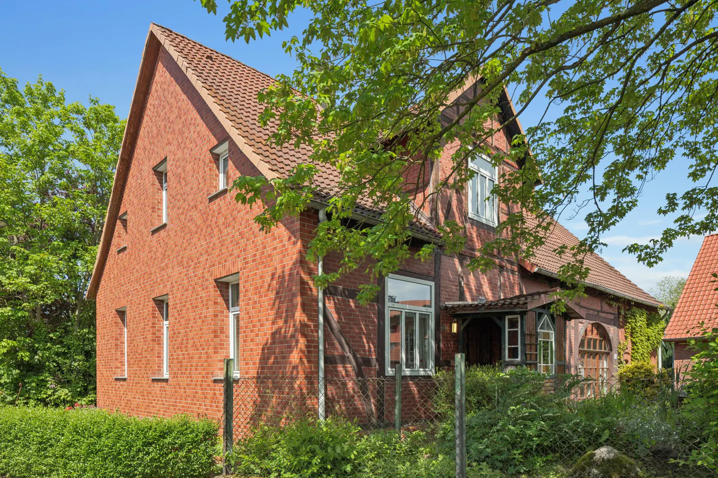 A two-story red brick house with a red tile roof, white windows, and green trees around it.
