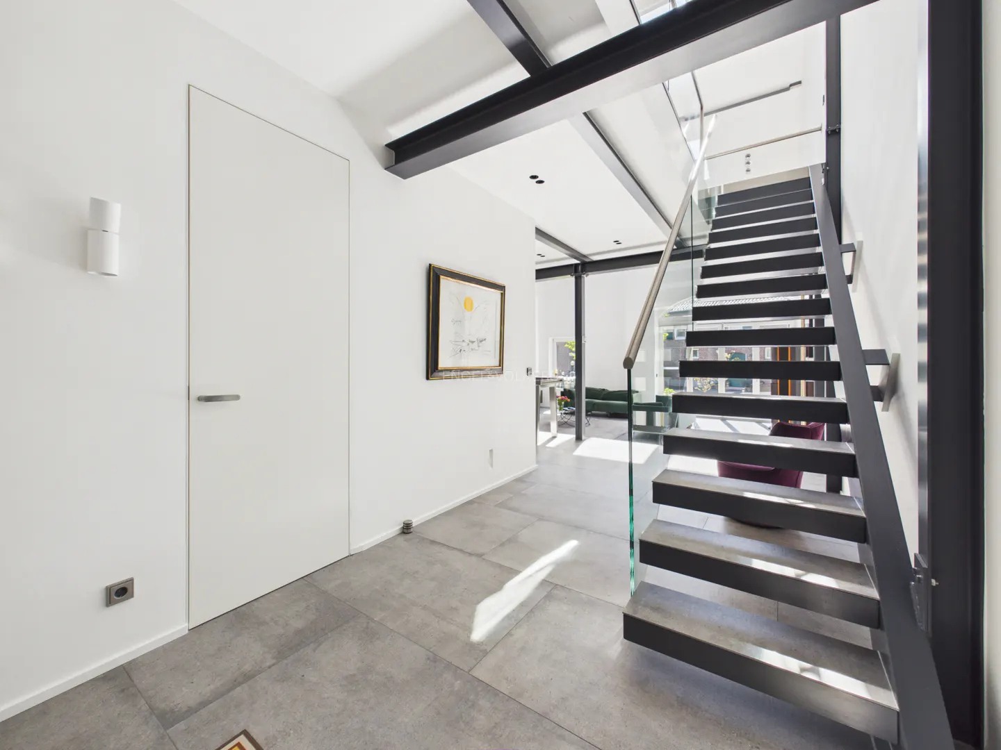 Bright, modern hallway with white walls, gray tile floor, and black staircase with glass railing. Artwork hangs on the wall.