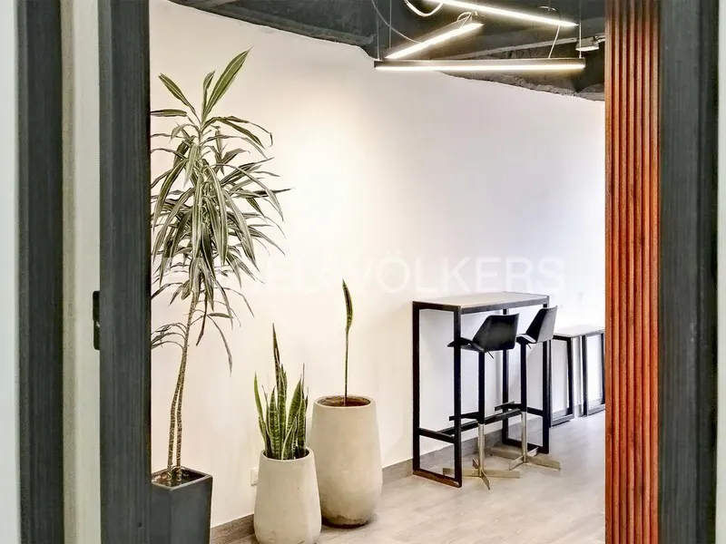 Interior view of a modern office space with plants, high tables, and black stools, seen through a doorway.