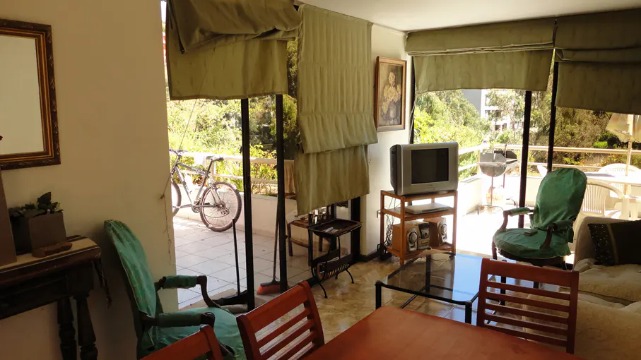A sunlit living room with a balcony view, featuring a TV, wooden furniture, and a bicycle parked outside.
