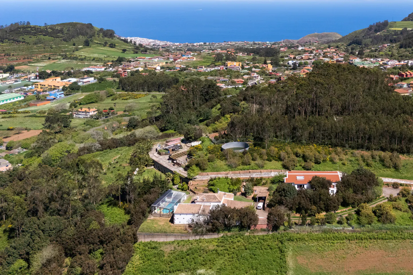 Aerial view of a white house with a pool, surrounded by green fields and trees, with a town and ocean in the background.