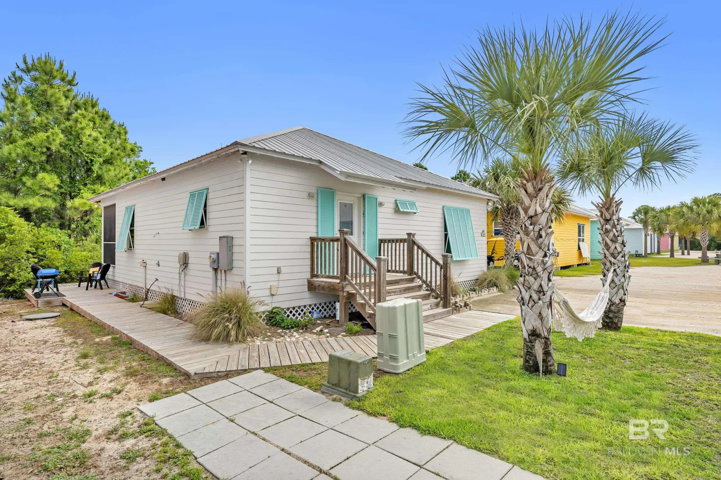 Exterior of a white beach house with turquoise shutters, a wooden deck, and palm trees with a hammock.