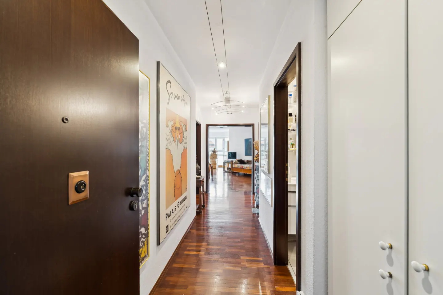 Hallway view with a dark wood door, posters on the white walls, and a shiny wood floor leading to a living room.