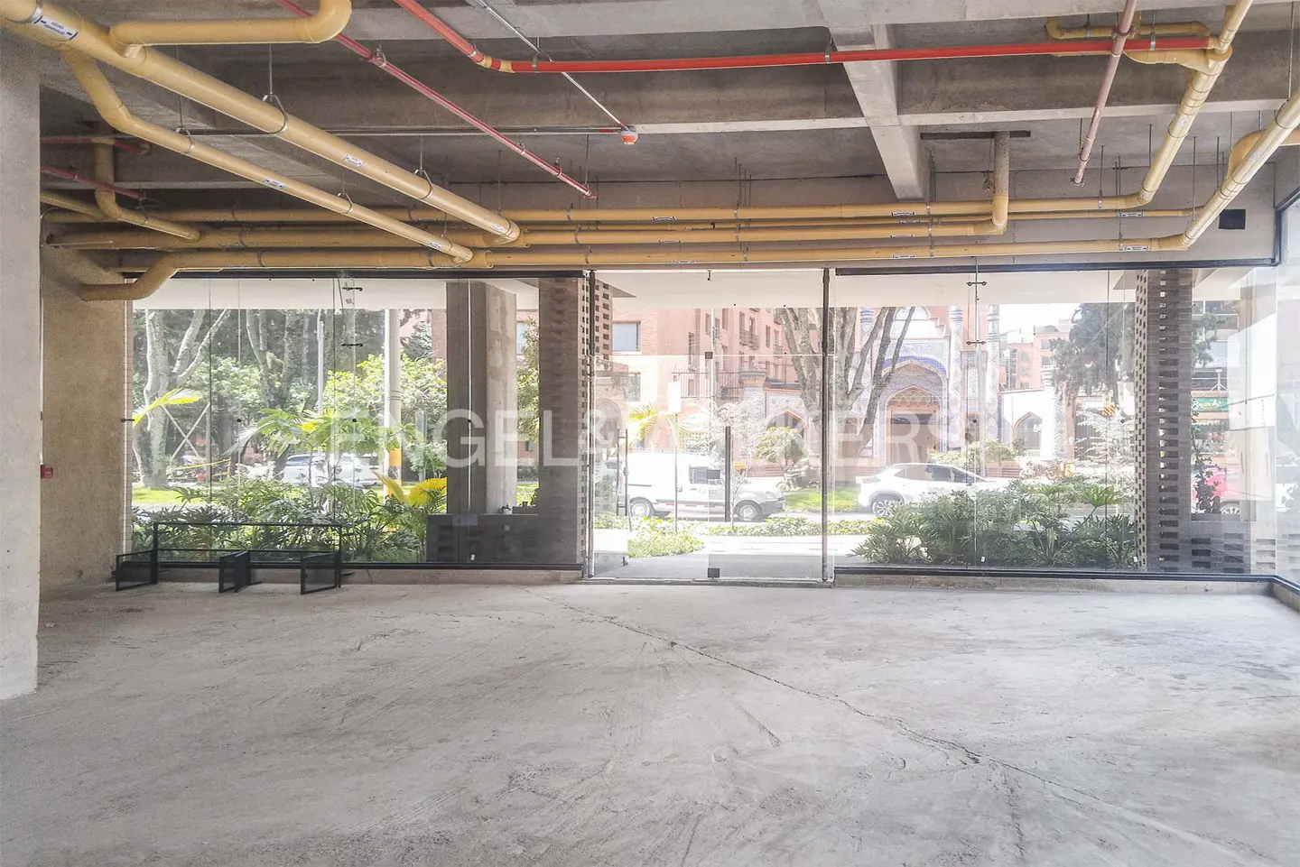 Unfinished commercial space with concrete floors and exposed ceiling pipes. Large glass windows offer a view of greenery and a building across the street.