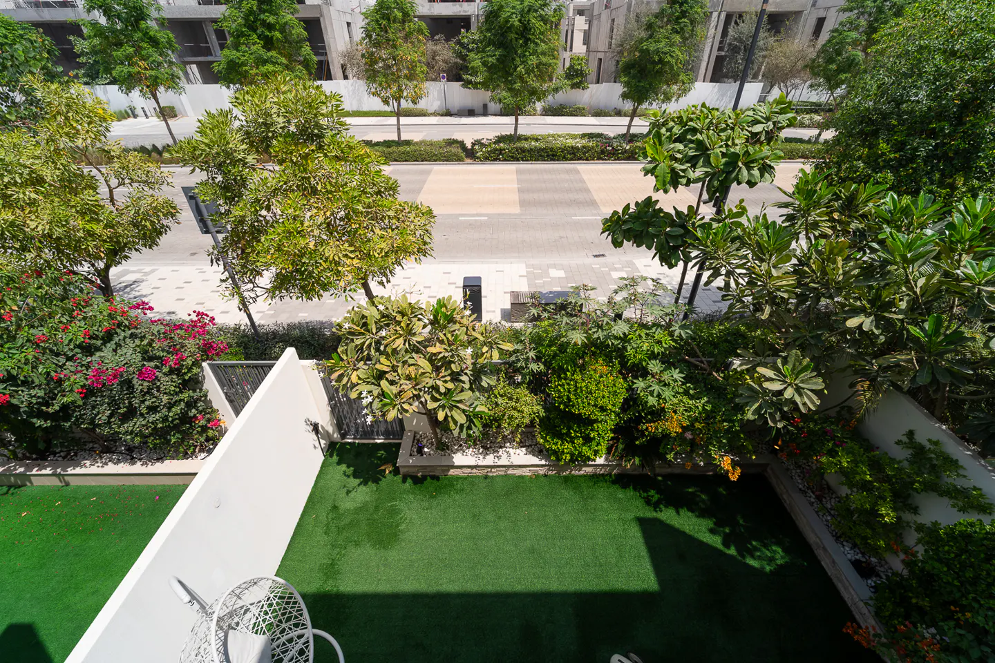 Aerial view of a green backyard with artificial grass, a white wall, and a white chair. Lush trees and a street are visible in the background.
