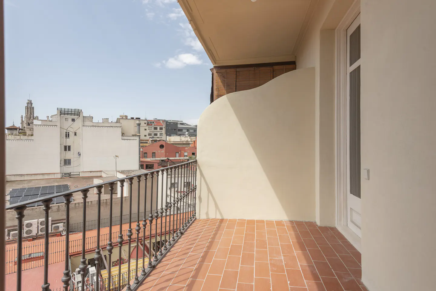 A balcony with red tile flooring and a black iron railing overlooks a cityscape under a blue sky. A white door is on the right.