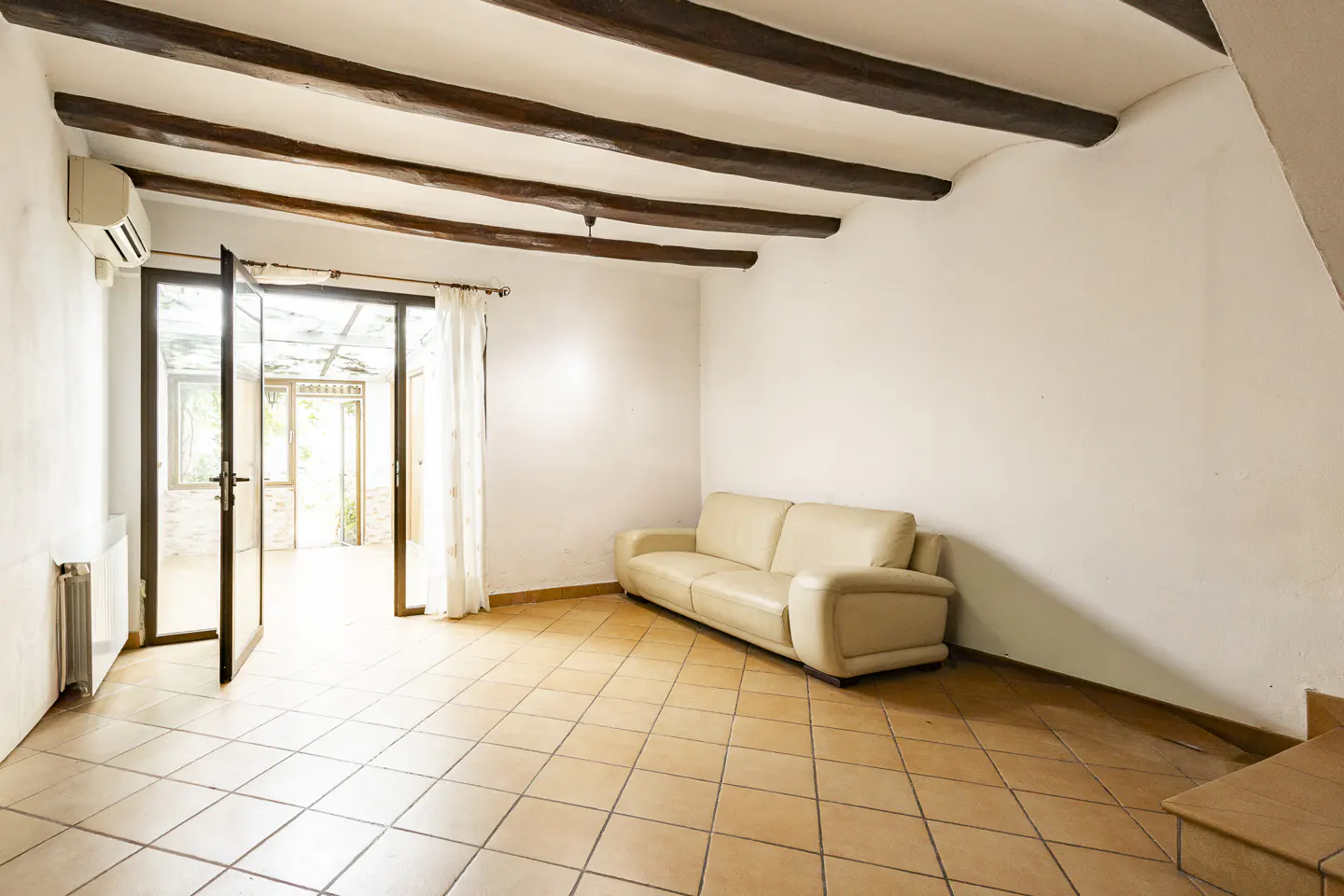 Bright living room with beige tile floor, white walls, and exposed dark wood beams. A beige sofa sits near open glass doors with sheer curtains.