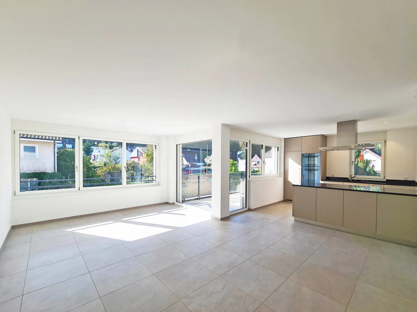 Bright, open-concept living space with tile floors, white walls, and large windows. A modern kitchen with beige cabinets is visible.