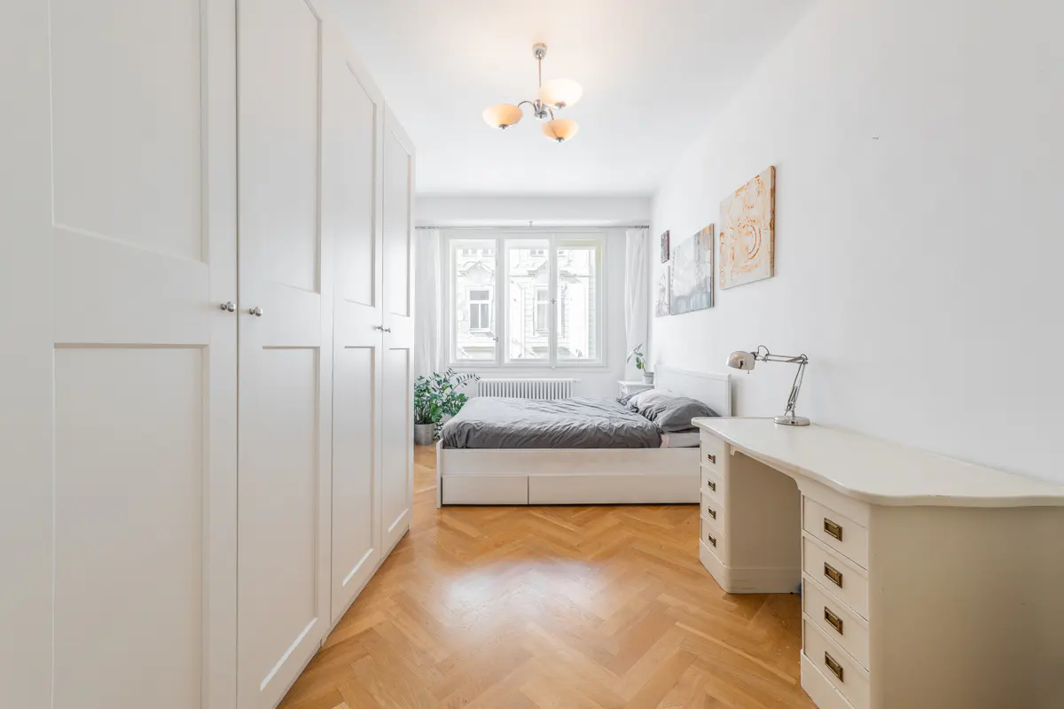 Bedroom with white walls, herringbone wood floor, bed, desk, and wardrobe. A window provides natural light.