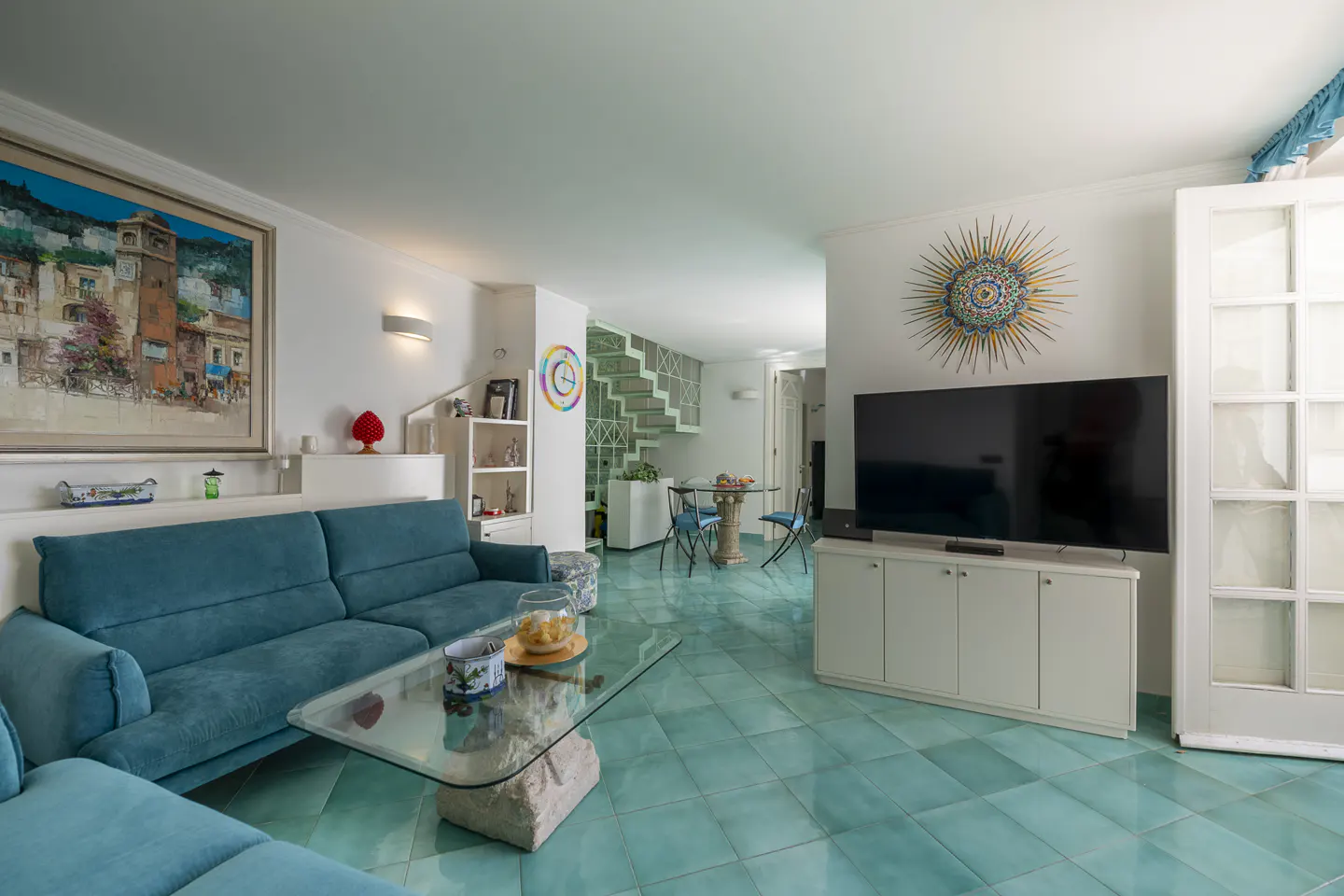 Bright living room with turquoise tile floor, teal sofa, glass table, and large TV. A painting and sunburst wall art add color.