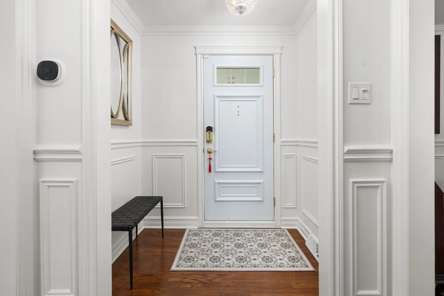 Bright foyer with white walls, wainscoting, and a white front door. A black bench sits to the left, and a patterned rug lies on the wood floor.