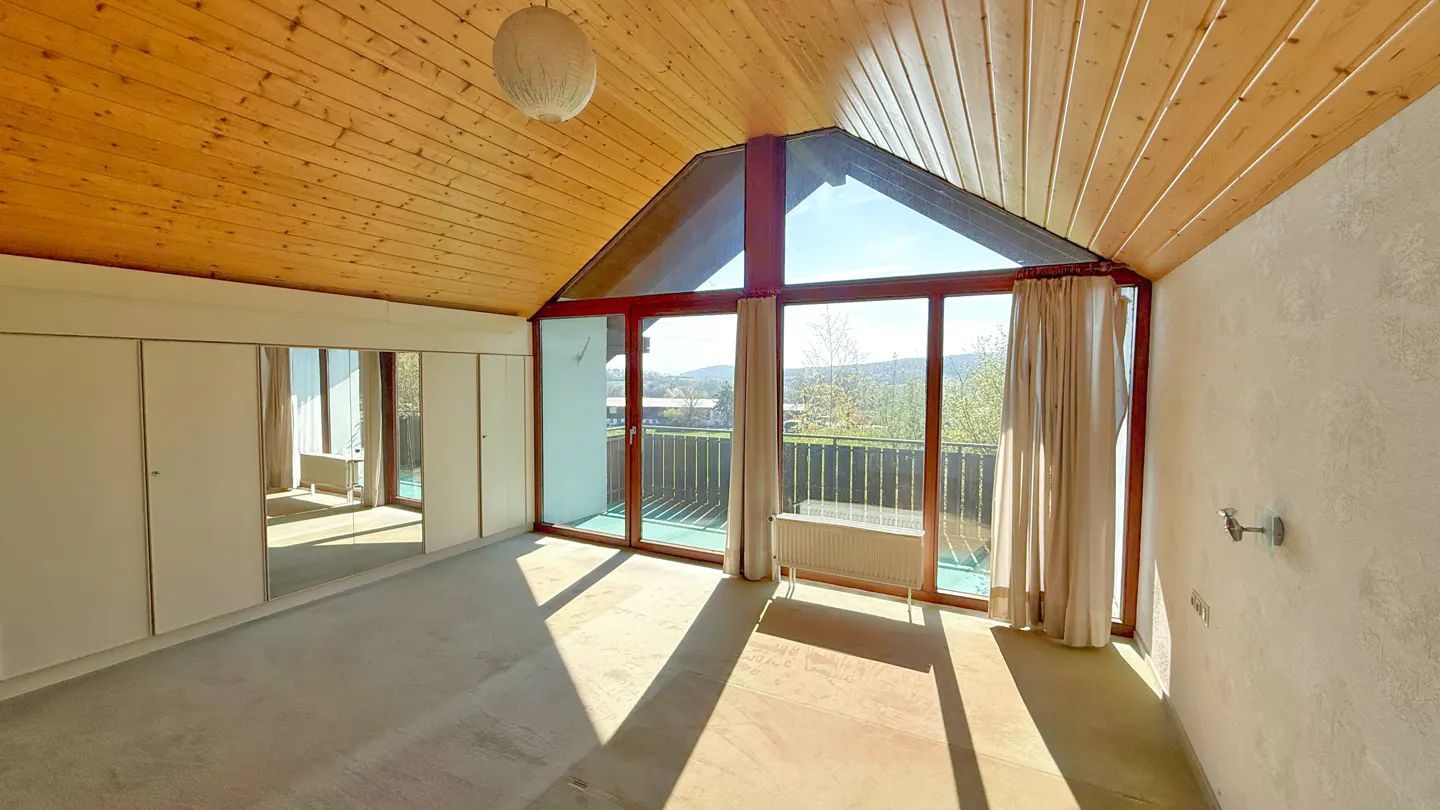 Bright room with wood ceiling, large windows, and balcony. White wardrobe with mirrored doors on the left. Curtains frame the view.