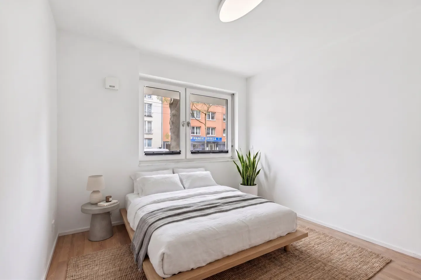 Bright bedroom with white walls, a wooden bed frame, white bedding, and a gray throw blanket. A window overlooks a city street.