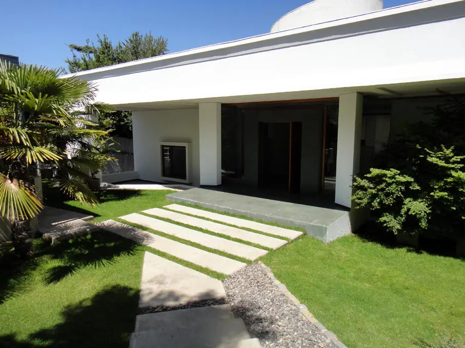 Modern white house exterior with a flat roof, green lawn, and stone pathway leading to the entrance. Palm tree on the left.