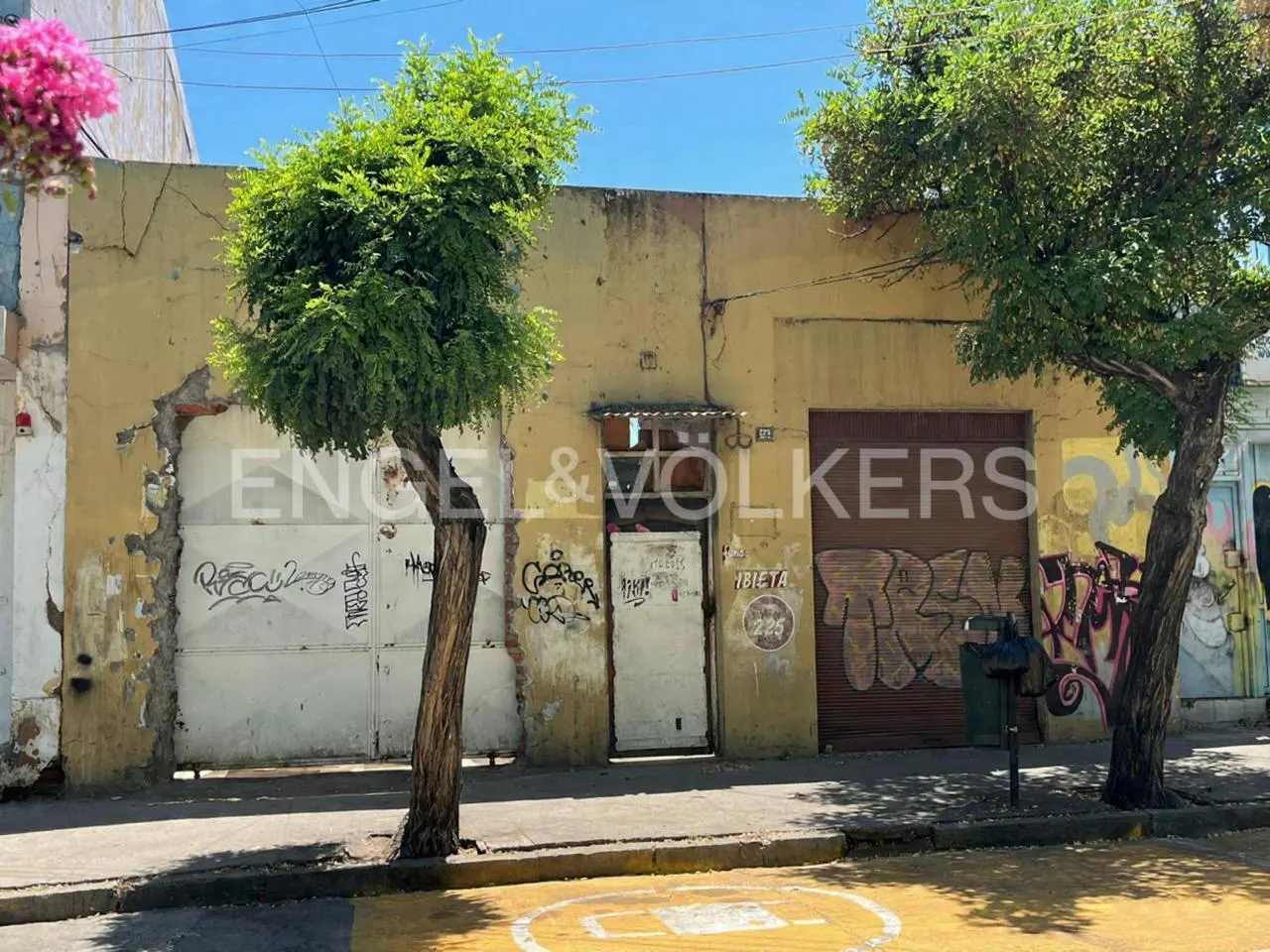 Exterior view of a yellow building with graffiti, trees, and an Engel & Volkers logo. The building has a door and roll-up garage doors.