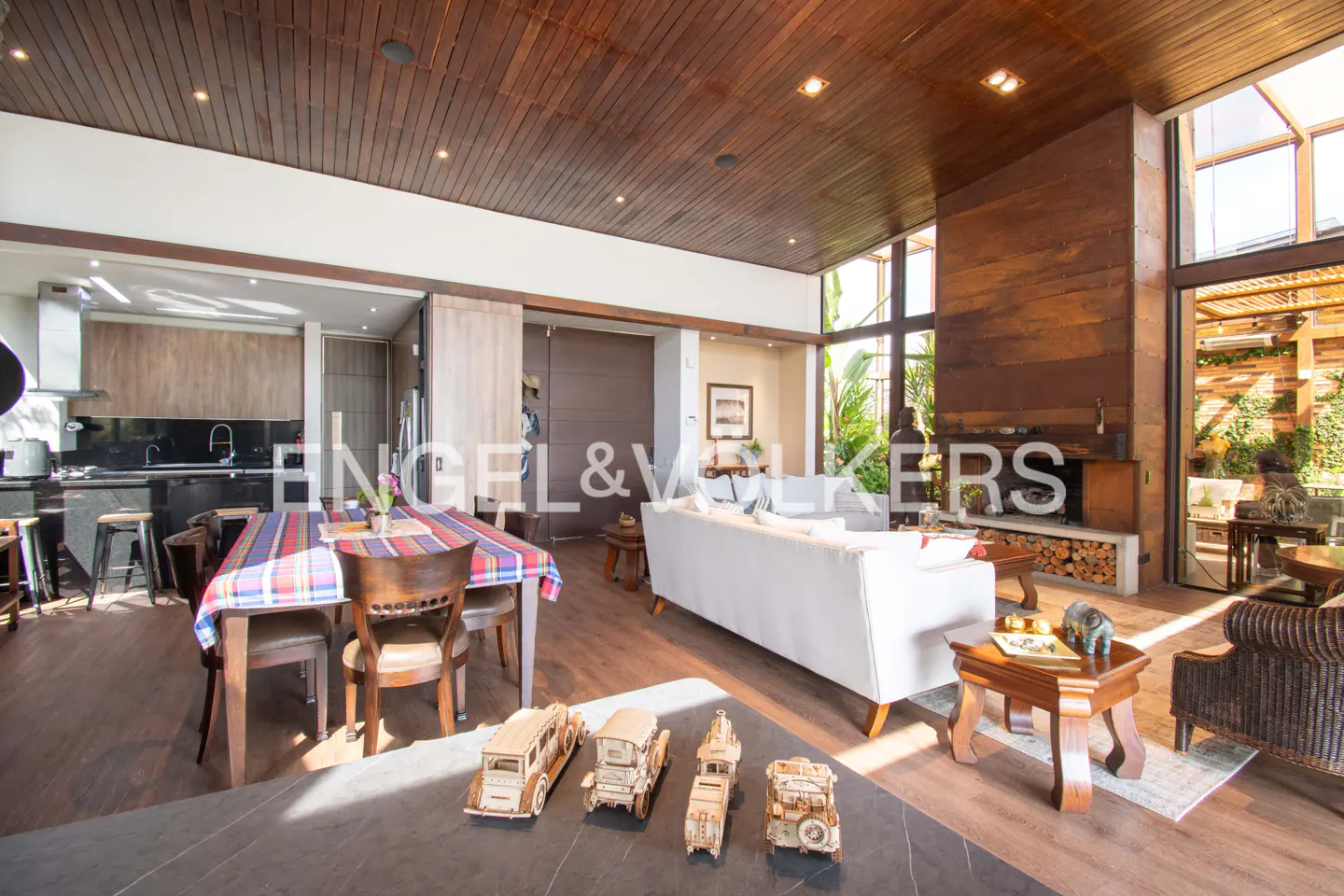 Open-concept living space with wood ceiling, kitchen, dining table with plaid cloth, white sofa, and fireplace.
