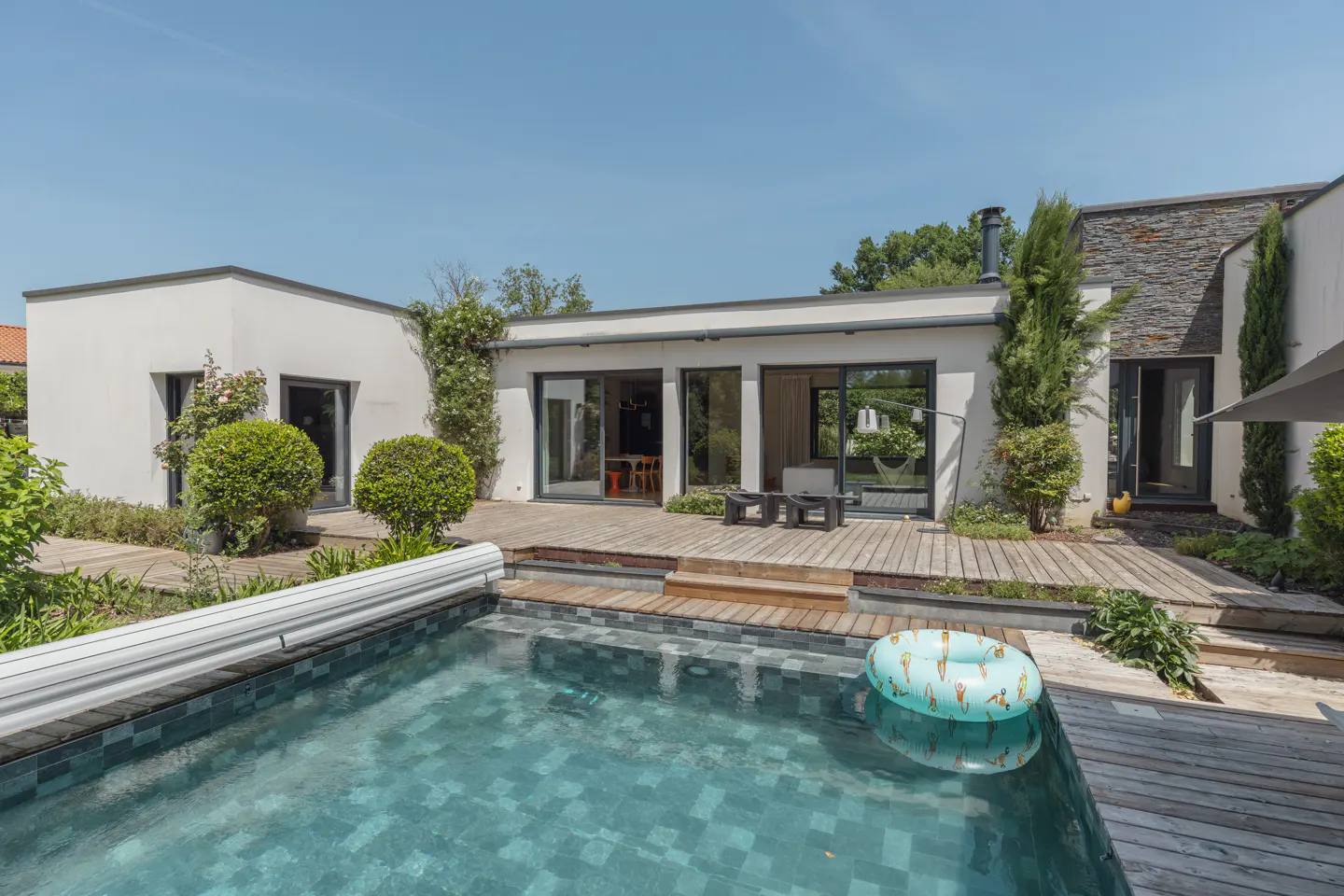 A modern white house with a pool and wooden deck on a sunny day. A blue inflatable ring floats in the pool.