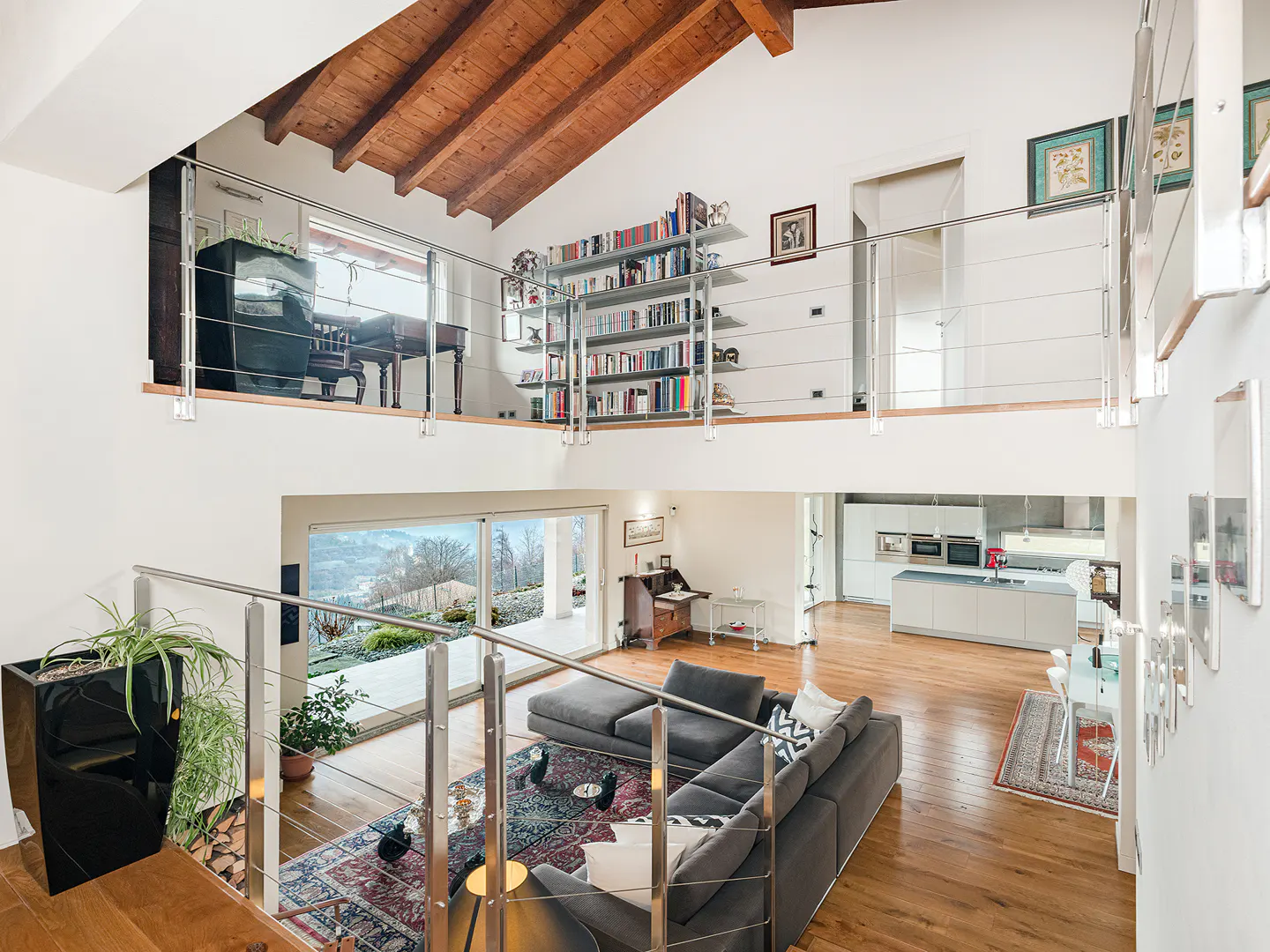 A modern, two-story living room with a gray sectional sofa, wood floors, and a view of the outside. A piano and bookshelf are on the second floor.
