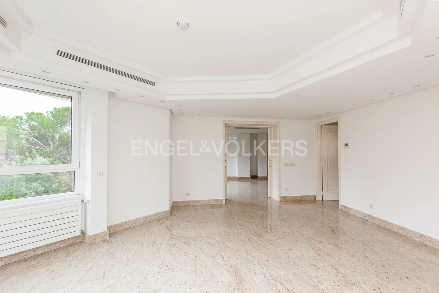 Bright, empty room with marble floors, white walls, and large window showing trees. Doors lead to other rooms. Engel & Völkers logo visible.