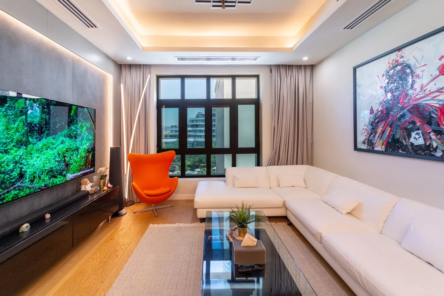 Living room with a white sectional sofa, orange chair, and a large TV displaying a green forest scene. A black framed window shows a city view.