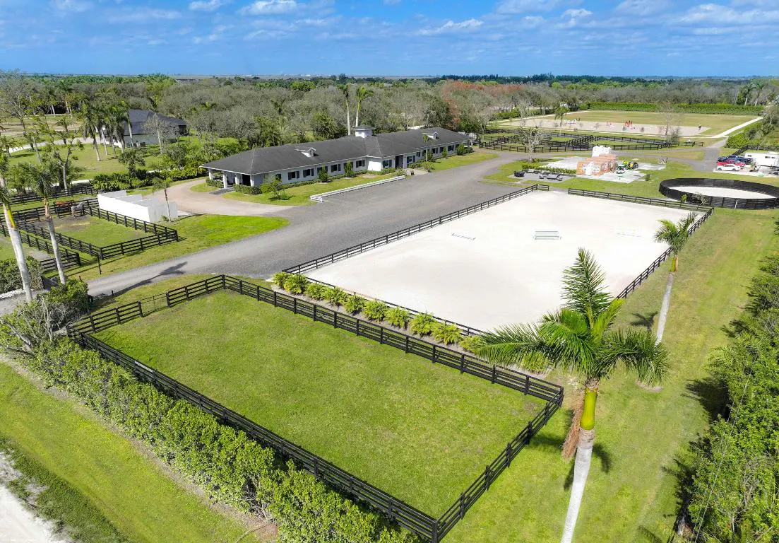 Aerial view of a horse farm with green pastures, a white riding arena, and a long white barn with a gray roof.