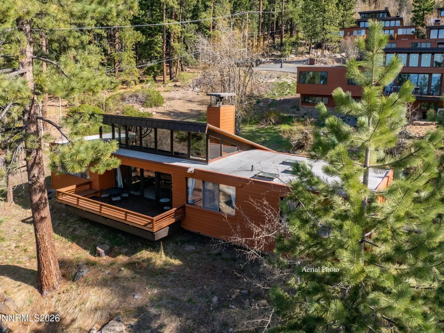 Aerial view of a modern, orange-toned house with a flat roof, surrounded by green trees. A wooden deck extends from the side.