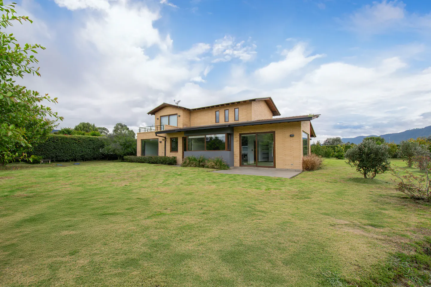 Two-story tan brick house with a balcony, large windows, and a green lawn under a cloudy sky.