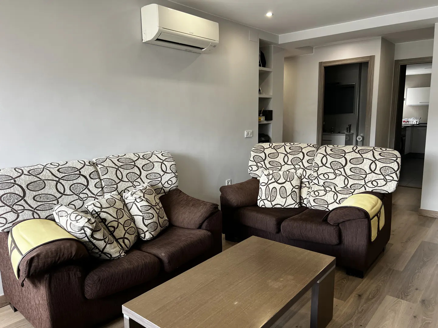 Living room with two brown sofas, patterned cushions, and a wooden coffee table. An air conditioner is mounted on the gray wall.