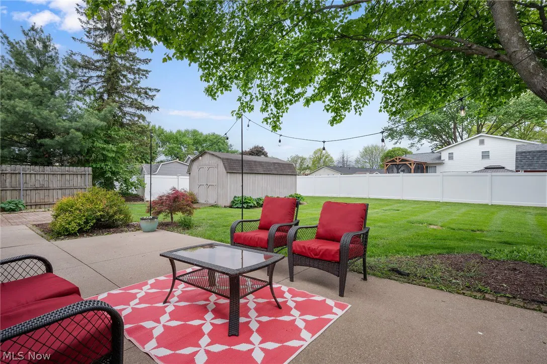 Backyard patio with red cushioned chairs, glass table on a red and white rug. Green lawn, white fence, and shed in the background.