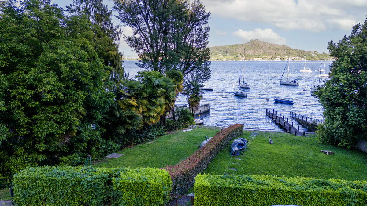 Lush green lawn slopes to a blue lake with sailboats and a dock. Trees frame the view, with a mountain in the distance.