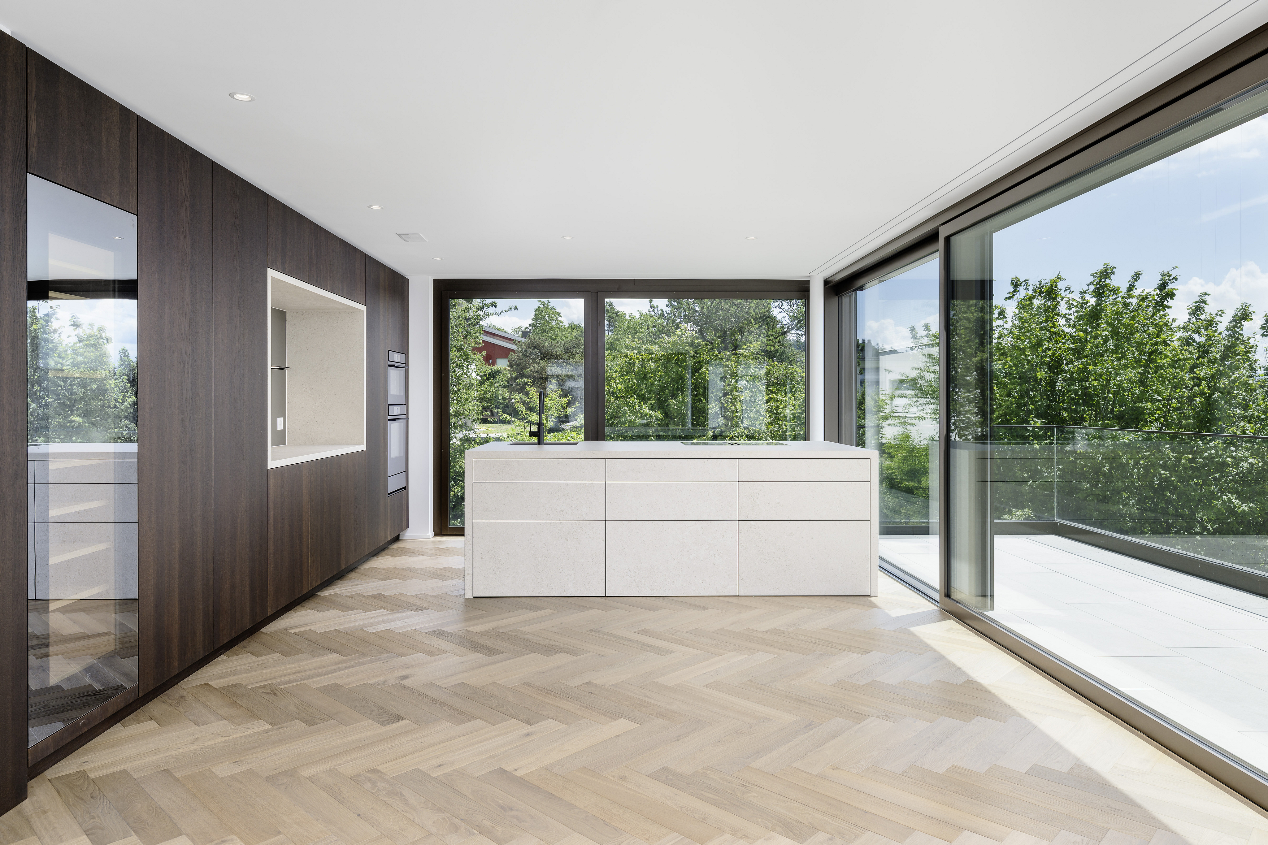 Modern kitchen with white island, dark wood cabinetry, herringbone wood floors, and large windows overlooking green trees.