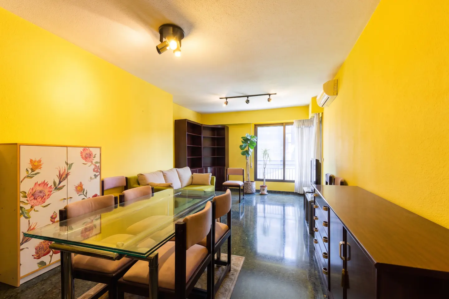 Bright yellow living room with a glass table, floral cabinet, and a green sofa. A large window lets in natural light.