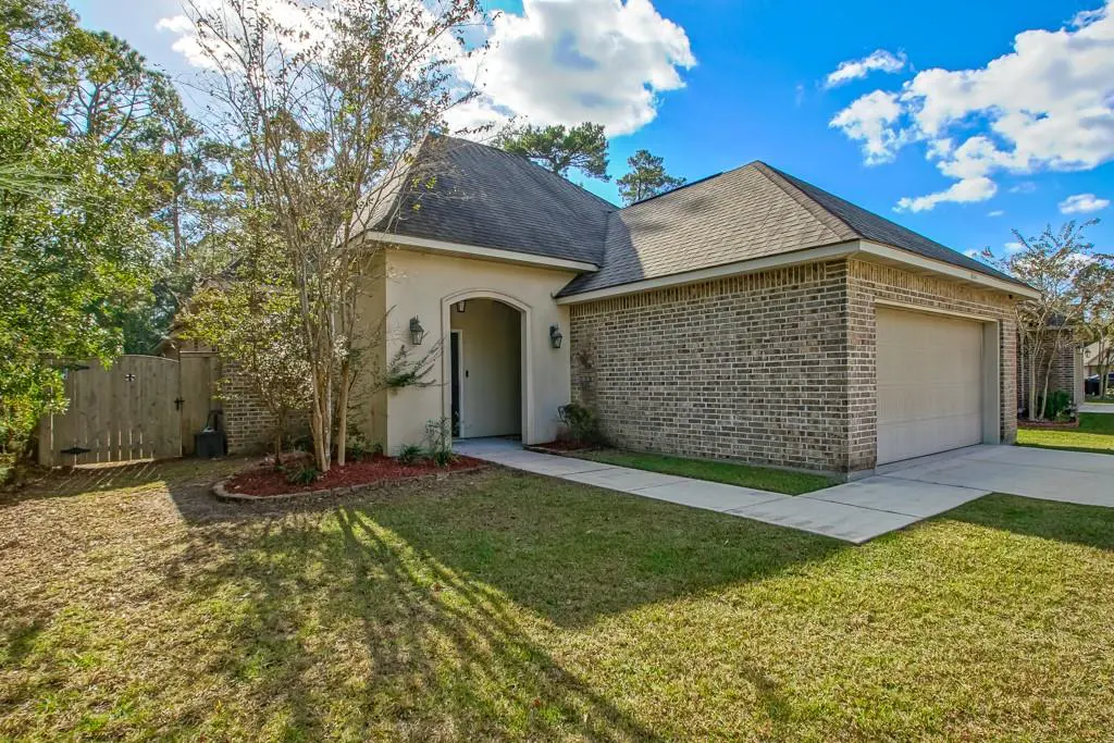 A single-story brick house with a gray roof, green lawn, and trees under a blue, cloudy sky.