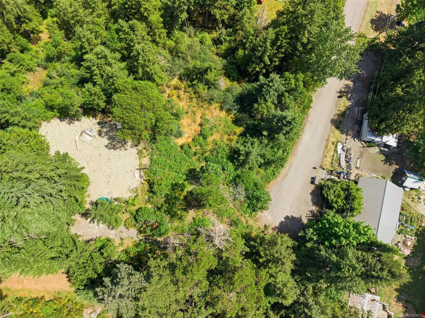Aerial view of a wooded lot with a cleared gravel area, adjacent to a paved road and a small building with a gray roof.