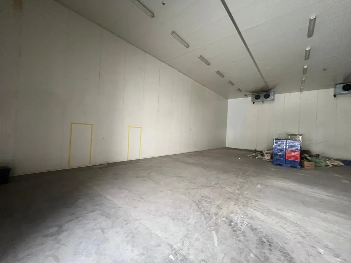 A large, empty warehouse with white walls, a concrete floor, and fluorescent lighting. Blue and red crates are stacked in the corner.
