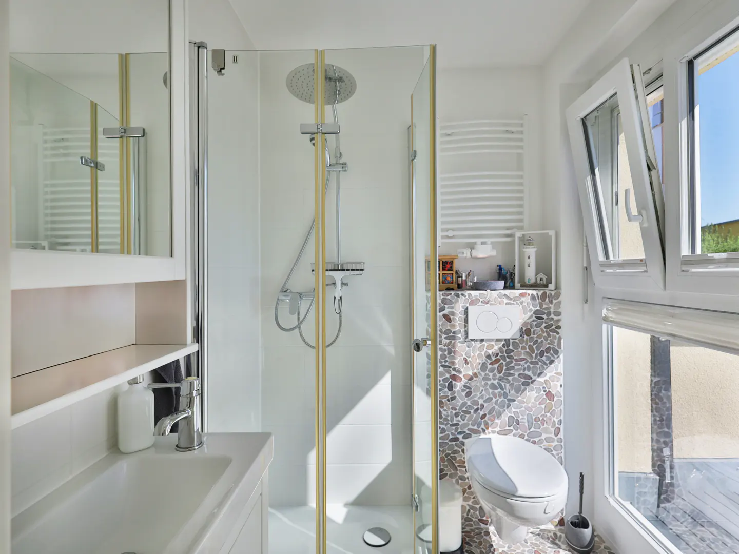Bright bathroom with glass shower, white sink, and pebble-tiled toilet area near an open window.