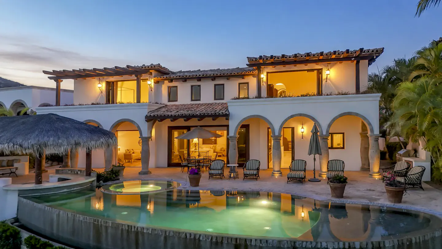 Exterior view of a two-story white villa with a pool, arches, and a thatched-roof cabana at dusk. Palm trees are visible.